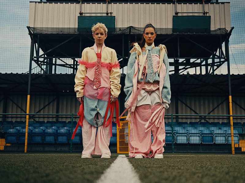 Two people dressed in elaborate colourful outfits pose on a football pitch.