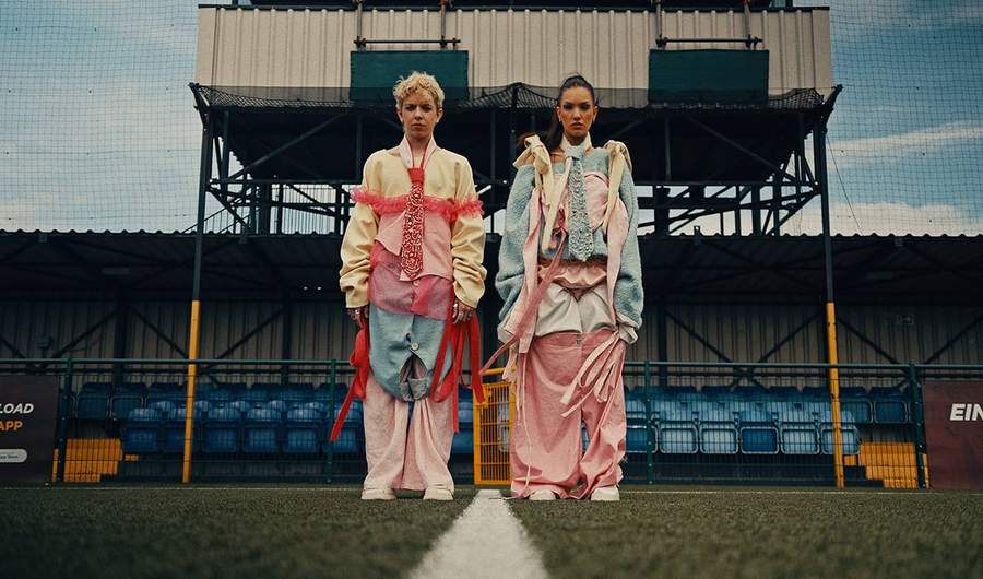 Two people dressed in elaborate colourful outfits pose on a football pitch.