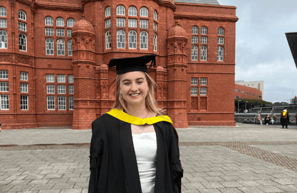 A young woman in a graduation cap and gown stands smiling in front of a historic red brick building. The gowns hood is yellow. The sky is overcast, and other people are seen in the background.