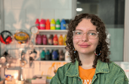A person with curly hair and glasses smiles in a colorful art studio. They wear a denim jacket over an orange shirt. In the background, shelves display vibrant paint bottles and art supplies.