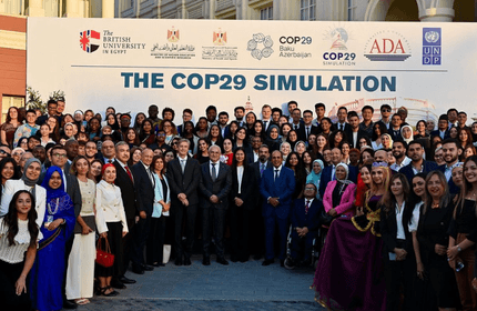 A large group of diverse people, dressed in formal and traditional attire, pose in front of a COP29 Simulation banner. The backdrop includes logos of various organizations. They appear to be participants in the event.