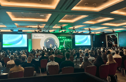 A large conference room filled with seated attendees facing a stage. The stage has a speaker, green lighting, and a screen displaying Deloittes logo and presentation slides. The ceiling features recessed lighting.