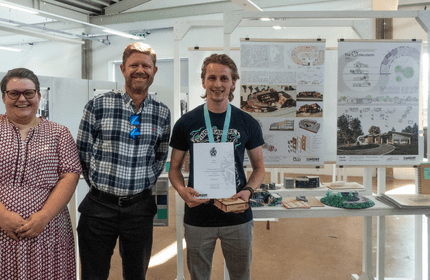 Three people stand smiling indoors near architectural posters. The person in the middle holds a certificate. The setting appears to be an exhibition or presentation space.