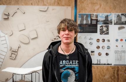 A person with shaggy hair stands in front of a wall displaying architectural designs and diagrams. They are wearing a black jacket and a shirt with a graphic design. The background features a 3D model and several project posters.