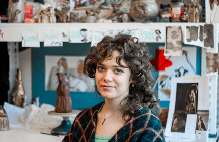 Student sits in front of artwork including pottery, sculptures and sketches