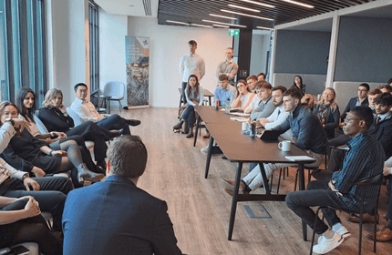 A group of people are seated in a modern conference room, attentively listening to a speaker at a meeting. The room has large windows and contemporary decor. Some attendees are taking notes, while others are engaged in the discussion.