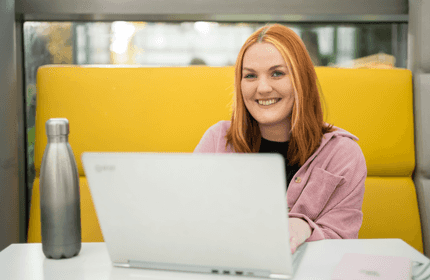 A person with red hair is sitting at a white table in a yellow booth, smiling and using a laptop. A silver water bottle is also on the table. The background shows large windows with a blurred outdoor view.