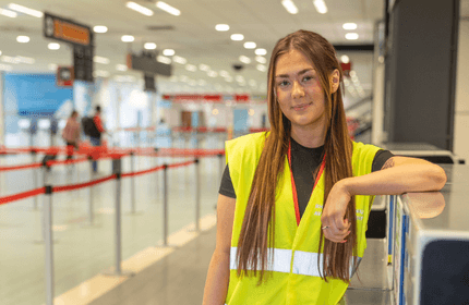 A person wearing a yellow safety vest stands inside an airport terminal, leaning on a counter, with several check-in counters and red ropes visible in the background. The terminal is well-lit and has a few people in the distance.