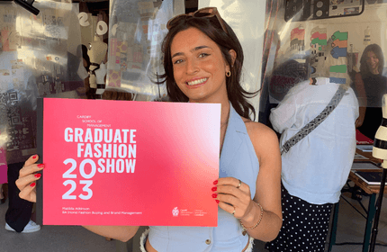 A woman smiles while holding a red sign that reads Graduate Fashion Show 2023, Cardiff School of Management. Shes standing in a room with people and mannequins in the background.