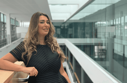 A person with long hair stands indoors, leaning on a railing in a modern building with large glass windows and a high ceiling. They are wearing a black dress, and there are reflections on the glass walls.