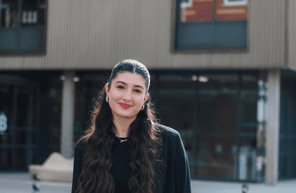 A person with long, wavy hair stands outdoors in front of a modern building. They are smiling and wearing a black top. The background features glass windows and a bench.