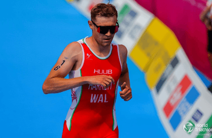 An athlete in a red triathlon suit is running with a blue track beneath. He wears sunglasses and has the number 18 on his left arm. The background shows colorful banners and the World Triathlon logo is visible at the bottom right.