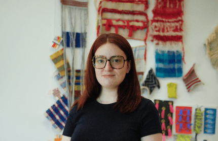 A woman with glasses stands in front of a colorful wall of textiles, showcasing various fabric patterns and textures.