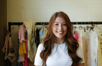 A woman with long auburn hair and a white shirt smiles at the camera, standing in front of a clothing rack with colorful garments.