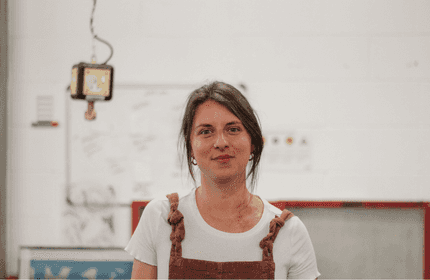 A woman wearing an apron stands in front of a textured wall, smiling and looking towards the camera.